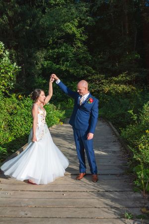 A couple in wedding attire dances together on a rustic wooden bridge surrounded by lush greenery.