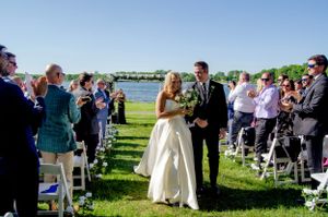 Outdoor wedding ceremony on a grassy lawn with guests seated in white chairs beside a lake.