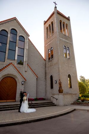 A wedding couple poses in front of a stone church with gothic architecture and bell tower.