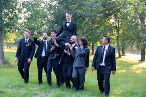 Groomsmen lift and carry a person in a playful pose outdoors on grassy lawn.
