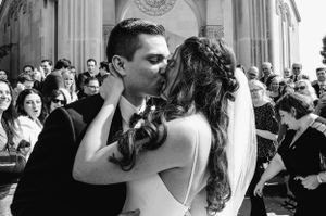A wedding couple shares their first kiss outside a grand church surrounded by guests.