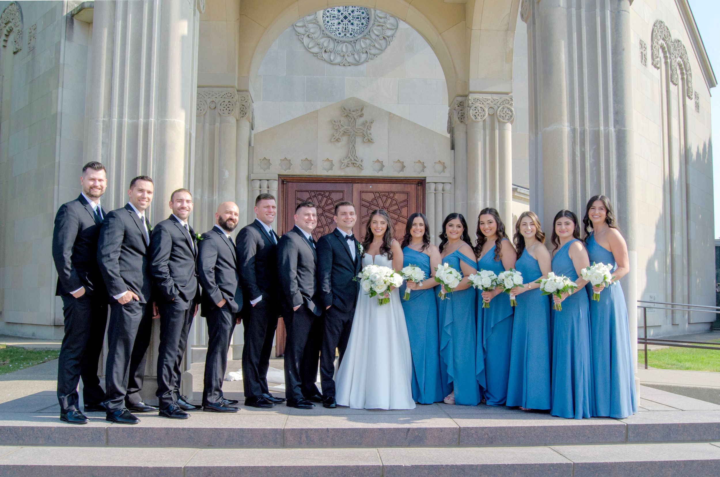 Wedding party in blue dresses and black suits poses under ornate archway architecture.