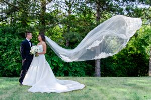Long flowing wedding veil catches wind as couple poses in outdoor garden setting.