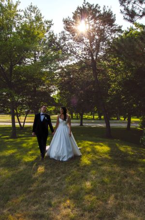 Newlyweds walking hand in hand through a sunlit park with trees.
