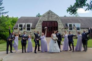 Wedding party in purple dresses and suits poses playfully outside stone building.