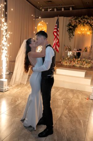A newlywed couple shares a romantic kiss on the dance floor with sparkling lights in the background.