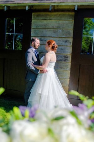 Bride and groom embracing by rustic wooden barn with purple flowers in foreground.