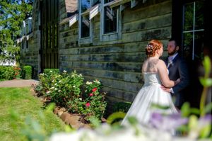 Wedding couple stands by weathered barn wall with blooming garden flowers.