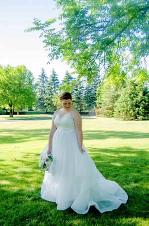 Bride in flowing white gown poses on sunny lawn with evergreen trees in background.