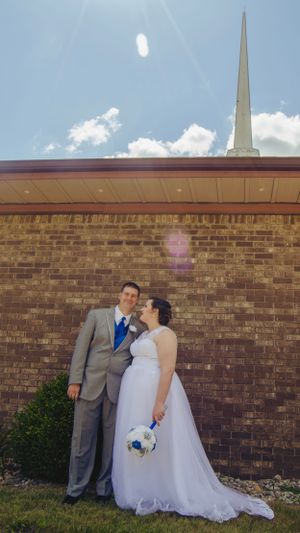 Wedding couple poses against brick church wall with white steeple in background.