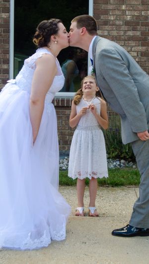 A sweet moment at a wedding with a flower girl reacting joyfully as the couple shares a kiss.