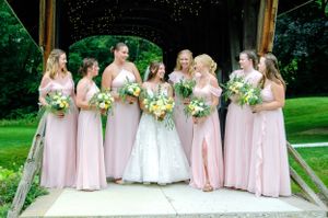 Bridal party in pink dresses poses with bride in white on a wooden bridge surrounded by greenery.