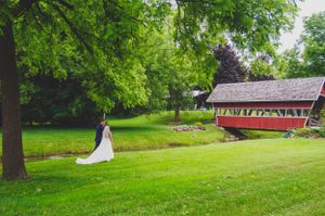 White wedding dress contrasts against red covered bridge and green landscape.