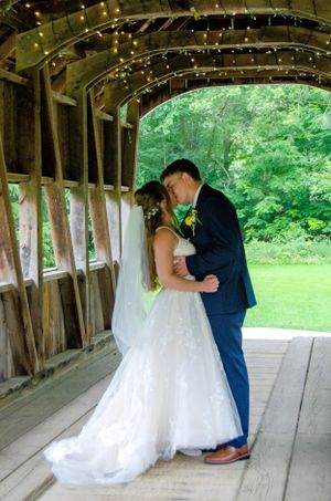 Couple shares intimate moment inside rustic covered bridge with string lights overhead.