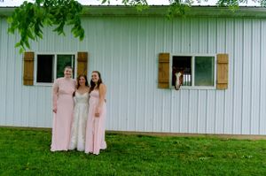 A group poses in front of a white barn with rustic wooden shutters wearing pink dresses.
