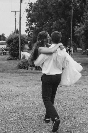 Groom carries bride in white dress while spinning on gravel path in black and white photo.