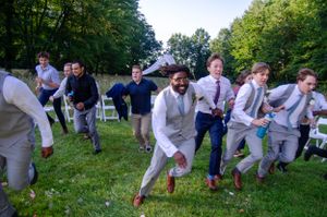 Wedding party runs and jumps playfully on grassy lawn during outdoor celebration.