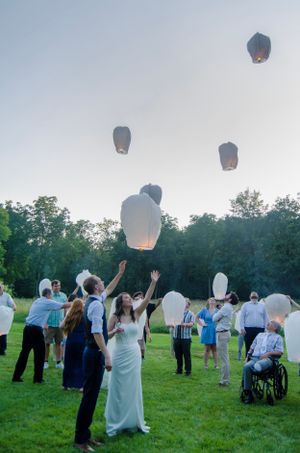 Wedding guests release white paper lanterns into twilight sky during outdoor reception.