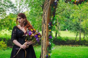 Person in black dress holding purple flowers in front of decorated garden arch.