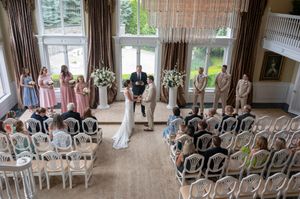 An overhead view of an elegant indoor wedding ceremony with rose-colored bridesmaids dresses and vintage white chairs.