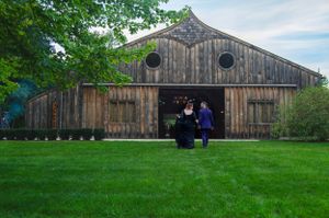 A pair in dark formal attire pose together in front of a weathered wooden barn with circular windows.