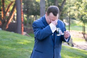Groom in blue suit wipes tears from eyes during emotional wedding moment.
