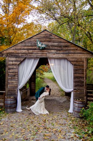 A decorated rustic barn entrance with white curtains and autumn foliage in the background.