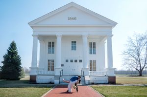 Couple sharing romantic dip kiss on brick path before historic 1846 white Greek Revival building.