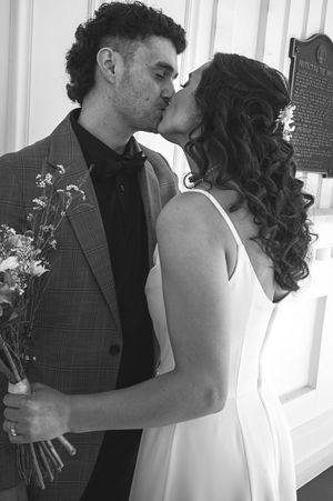 Black and white photo of a couple sharing a kiss, groom holding wildflowers, bride in white dress, wedding day moment.