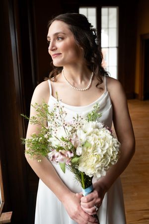 Bride in white satin gown holding white floral bouquet with pearl necklace, gazing softly to the side in elegant indoor setting.