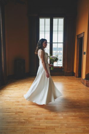 Bride in white gown twirling in sunlit wooden hall, holding bouquet, elegant wedding dress photography.