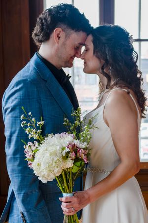 A bride and groom share an intimate moment, touching foreheads while she holds a bouquet of white hydrangeas and pink flowers.
