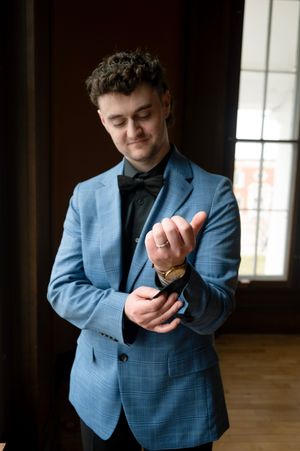 A groom in a blue plaid suit and black bow tie adjusts his gold cufflinks while wearing a wedding ring.