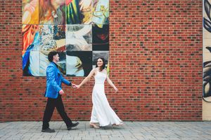Bride in white gown and groom in blue suit hold hands walking past a colorful brick wall mural.