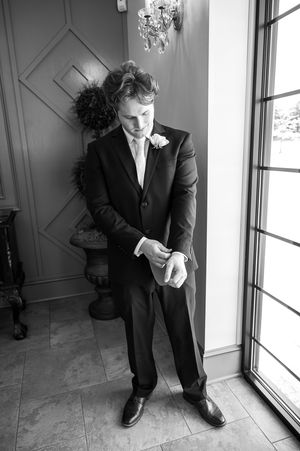 Black and white photo of a groom in a dark suit adjusting his cufflinks near a window before his wedding ceremony.