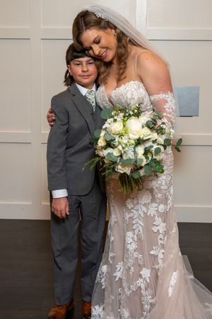 A bride in a delicate lace gown shares a sweet moment with a young ring bearer in a gray suit.