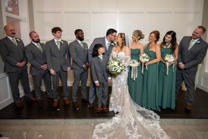 Wedding party in gray suits and emerald green dresses pose together for formal group photo with elegant decor.