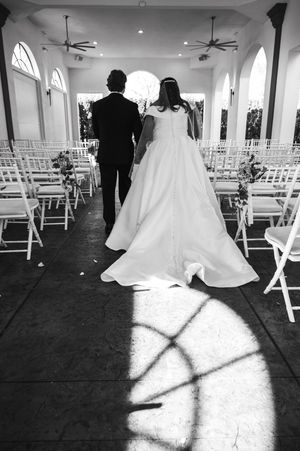 Black and white photo of bride and groom walking down aisle in elegant chapel with white chairs and arched windows.