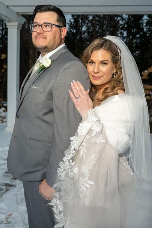 A winter wedding couple poses together in formal attire against a snowy outdoor backdrop with white columns.