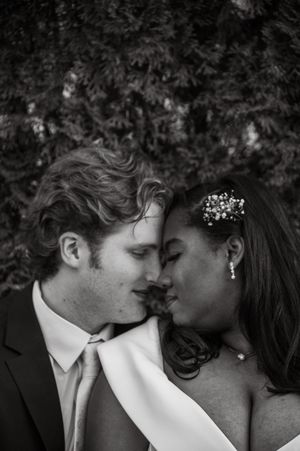 Black and white wedding photo of a couple in formal attire sharing an intimate moment, foreheads touching outdoors.