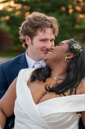 A joyful interracial couple shares a romantic kiss outdoors, dressed in wedding attire, with lush greenery in the background.