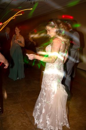 People dancing at a wedding reception with colorful lights and a bride in a white lace dress on the dance floor.