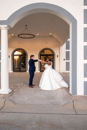 Bride in white ballgown and groom in navy suit dancing under elegant arched portico at wedding venue.