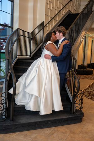 Bride and groom share a romantic kiss on an elegant staircase, bride in white gown, groom in navy suit.