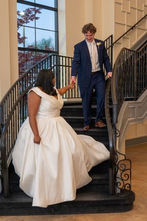 A bride in a white gown holds hands with a groom in a navy suit as they descend an elegant staircase.