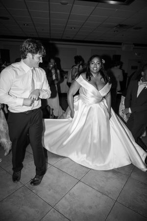 Black and white photo of a bride in a flowing gown dancing joyfully with a man in a dress shirt at a wedding reception.