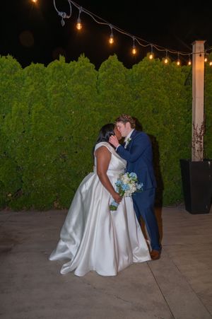 Bride and groom share a kiss outdoors at night, surrounded by string lights and a lush green hedge backdrop.