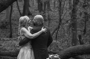 Black and white photo of a couple sharing a kiss in a forest, dressed in wedding attire.