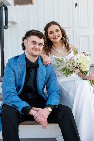 A smiling bride in white dress holding wildflowers poses with groom in blue plaid suit on church steps.