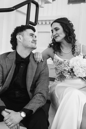 Black and white photo of a bride and groom sitting together, she holds a bouquet, both smiling at each other.